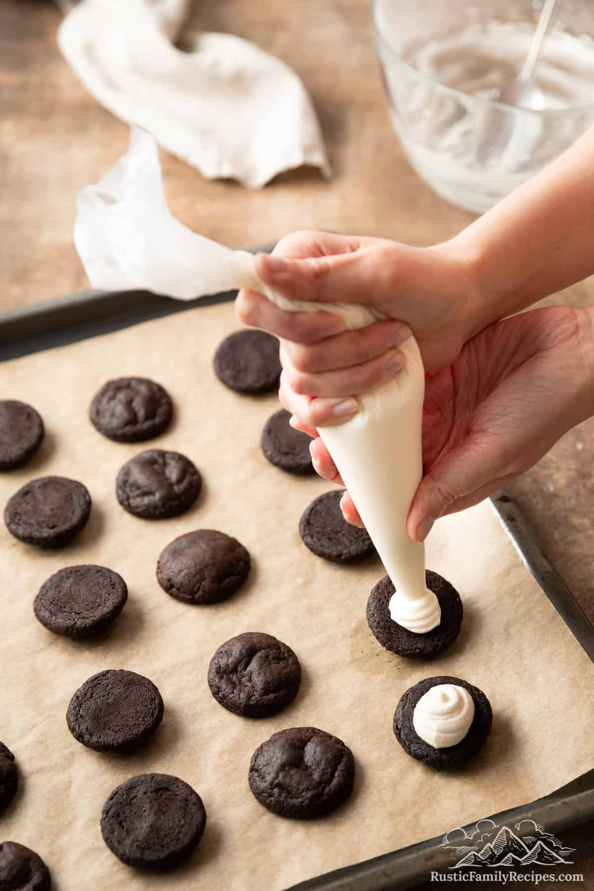 Hands use a piping bag to pipe vanilla filling onto chocolate cookie halves on a baking sheet.
