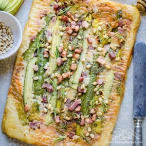 Top view of a zucchini tart topped with bacon, zucchini flowers and pine nuts.