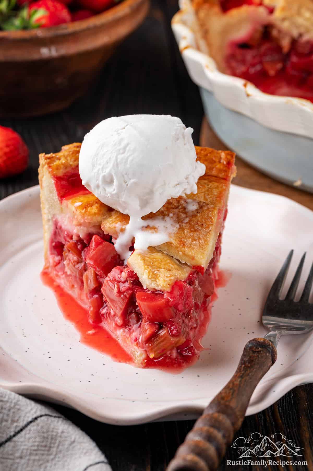 A slice of strawberry rhubarb pie facing forward on a white plate next to a fork, topped with whipped cream.