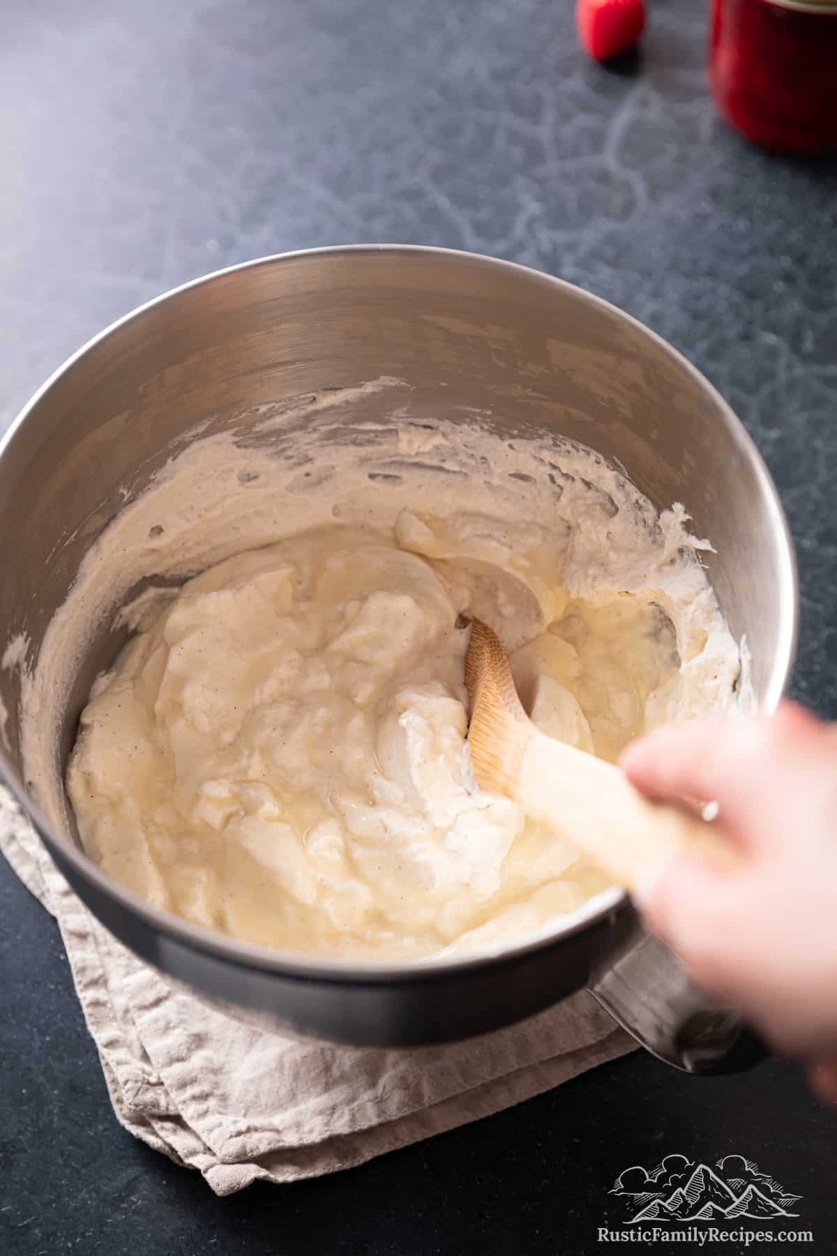 A wooden spatula is used to fold condensed milk into whipped cream in the bowl of a stand mixer.
