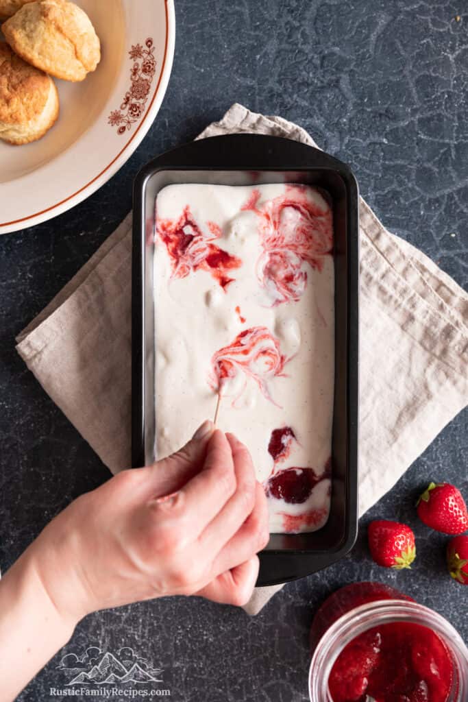 A hand uses a toothpick to swirl strawberry sauce into an ice cream base in a loaf pan.