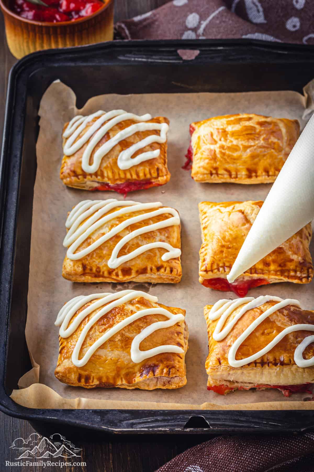 A piping bag is used to pipe squiggles of cream cheese frosting on top of baked strawberry toaster strudels on a baking sheet.