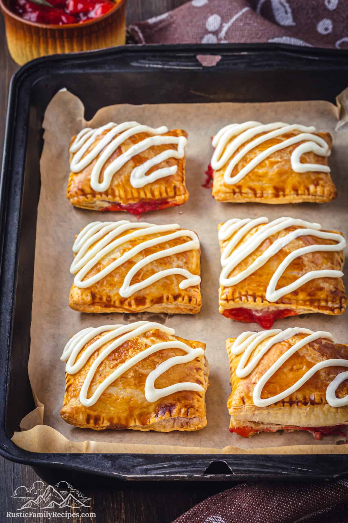 Baked strawberry toaster strudels on a baking sheet topped with squiggles of cream cheese frosting.