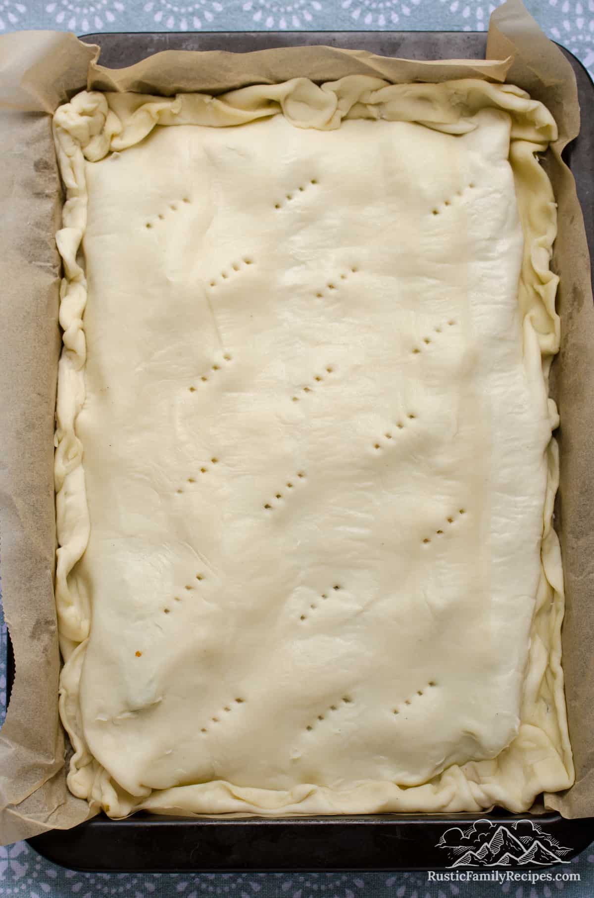 The topping ingredients for zucchini tart in a large rectangular parchment-lined baking pan, topped with pastry dough with fork pricks in the surface.