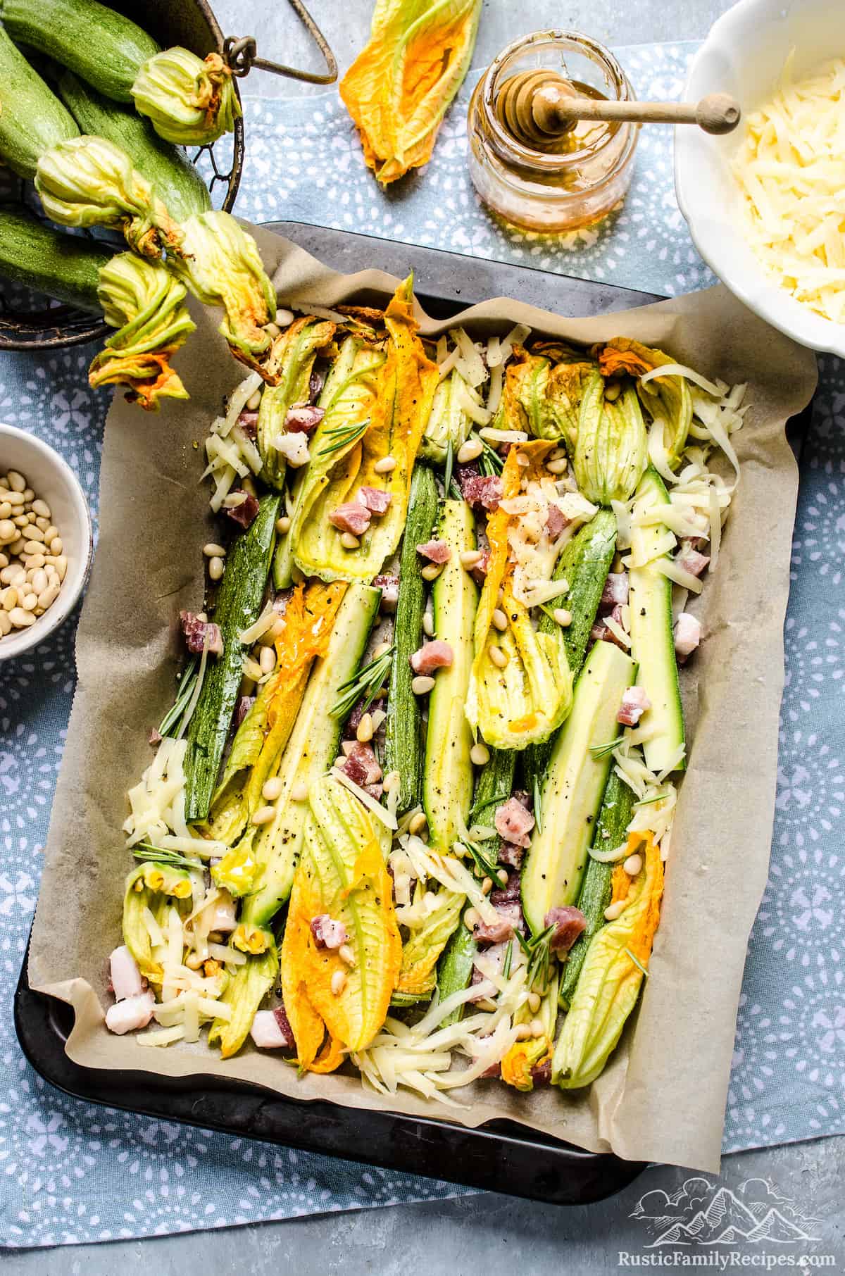 A large rectangular parchment-lined baking pan filled with zucchini strips, zucchini flowers, bacon and pine nuts, surrounded by small bowls holding the ingredients.