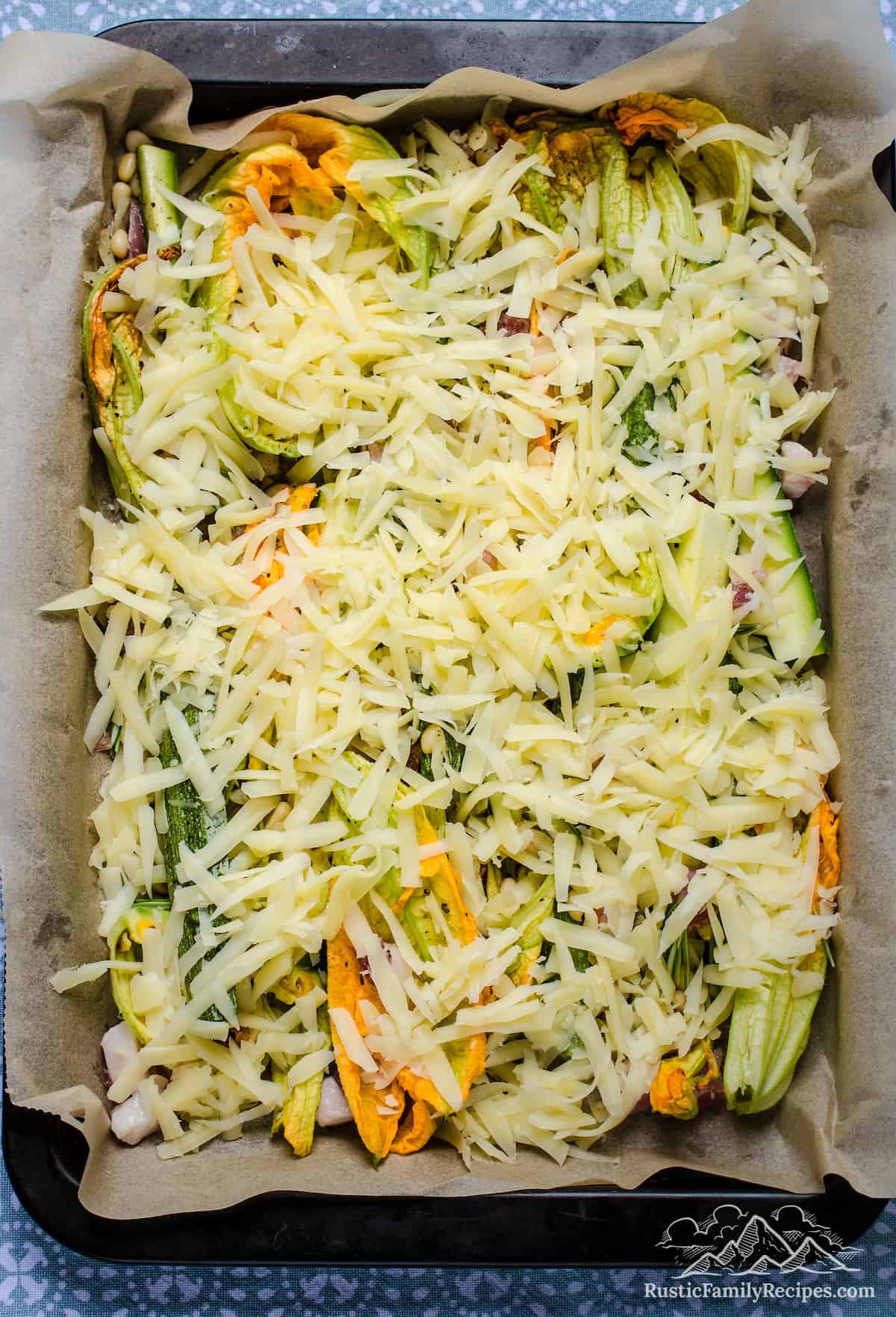 The topping ingredients for zucchini tart in a large rectangular parchment-lined baking pan, topped with a layer of shredded cheese.