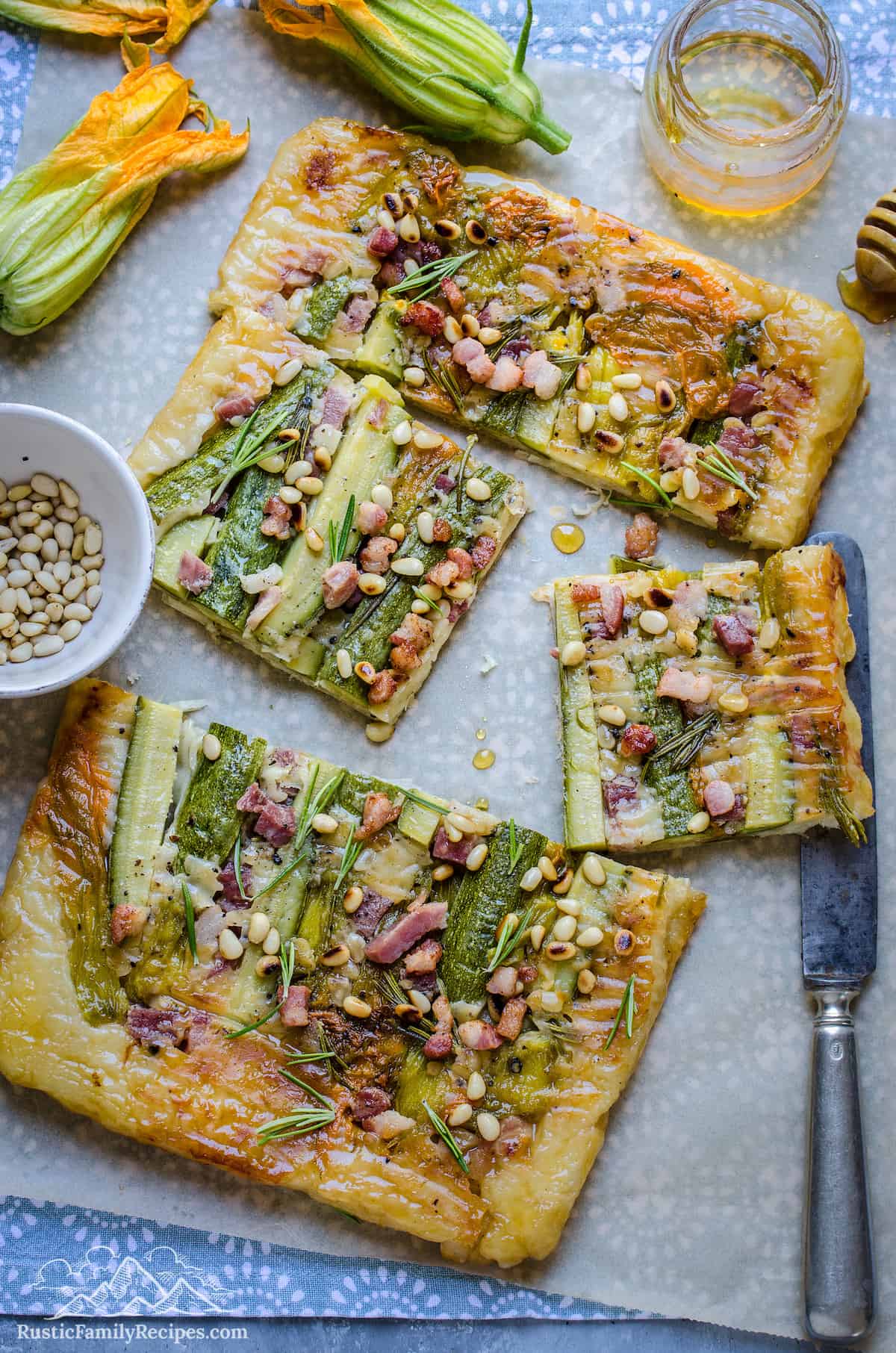 A zucchini tart cut into slices on top of a sheet of parchment paper, surrounded by a bowl of pine nuts and zucchini flowers.