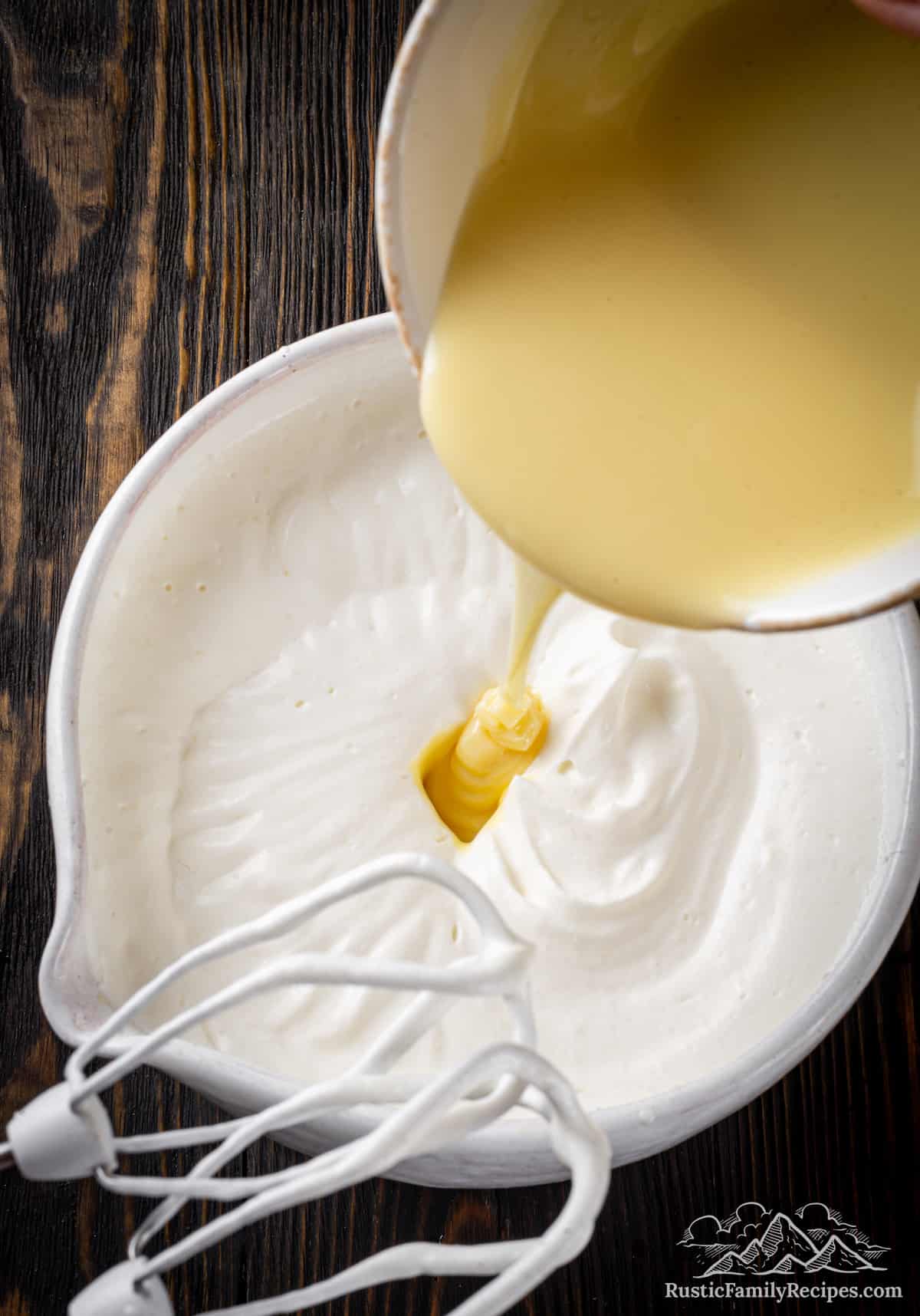 Condensed milk is poured into a bowl of whipped cream, next to a mixer's whisk attachment.