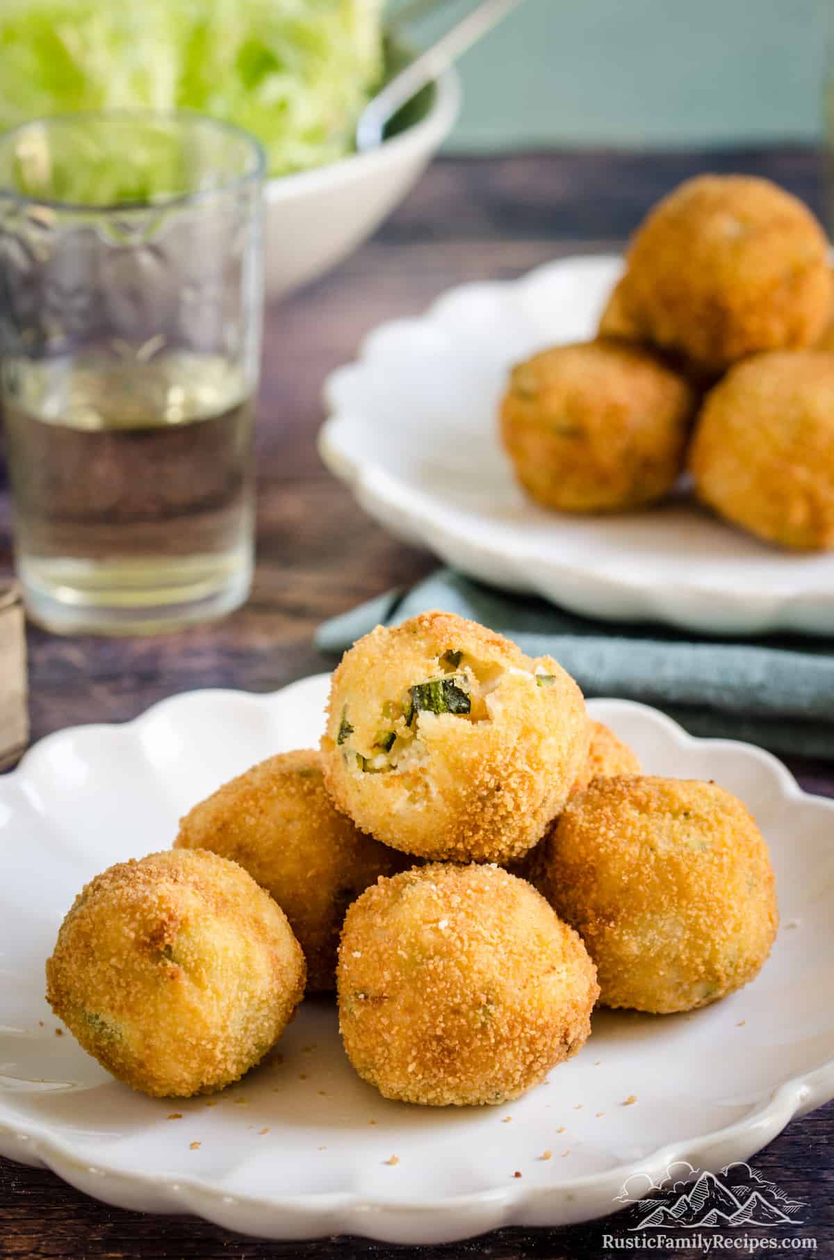 A pile of golden fried arancini with zucchini and goat cheese on a white plate, with a green salad and more arancini in the background.
