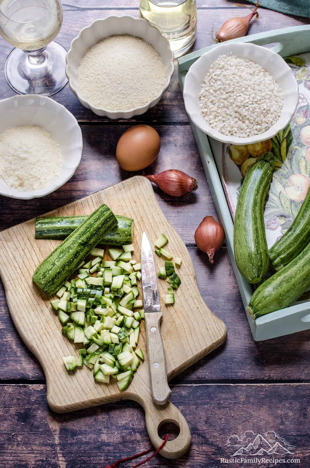 Diced zucchini on a wooden cutting board surrounded by the ingredients for homemade arancini.