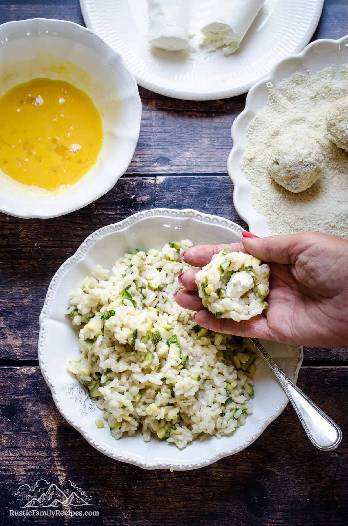 A hand holding a portion of cooked risotto with a piece of goat cheese in the center, over a bowl of zucchini risotto surrounded with bowls filled with coating ingredients.