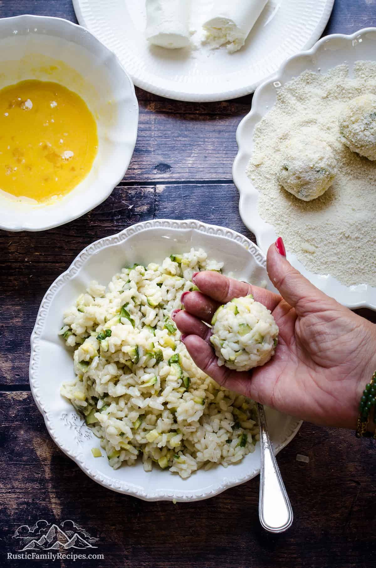 A hand holding a rice ball over a bowl of cooked zucchini risotto, surrounded by bowls filled with coating ingredients.