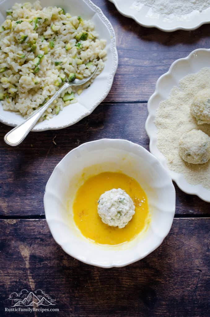 A risotto ball sits in a dish filled with egg to coat, next to a bowl of zucchini risotto.
