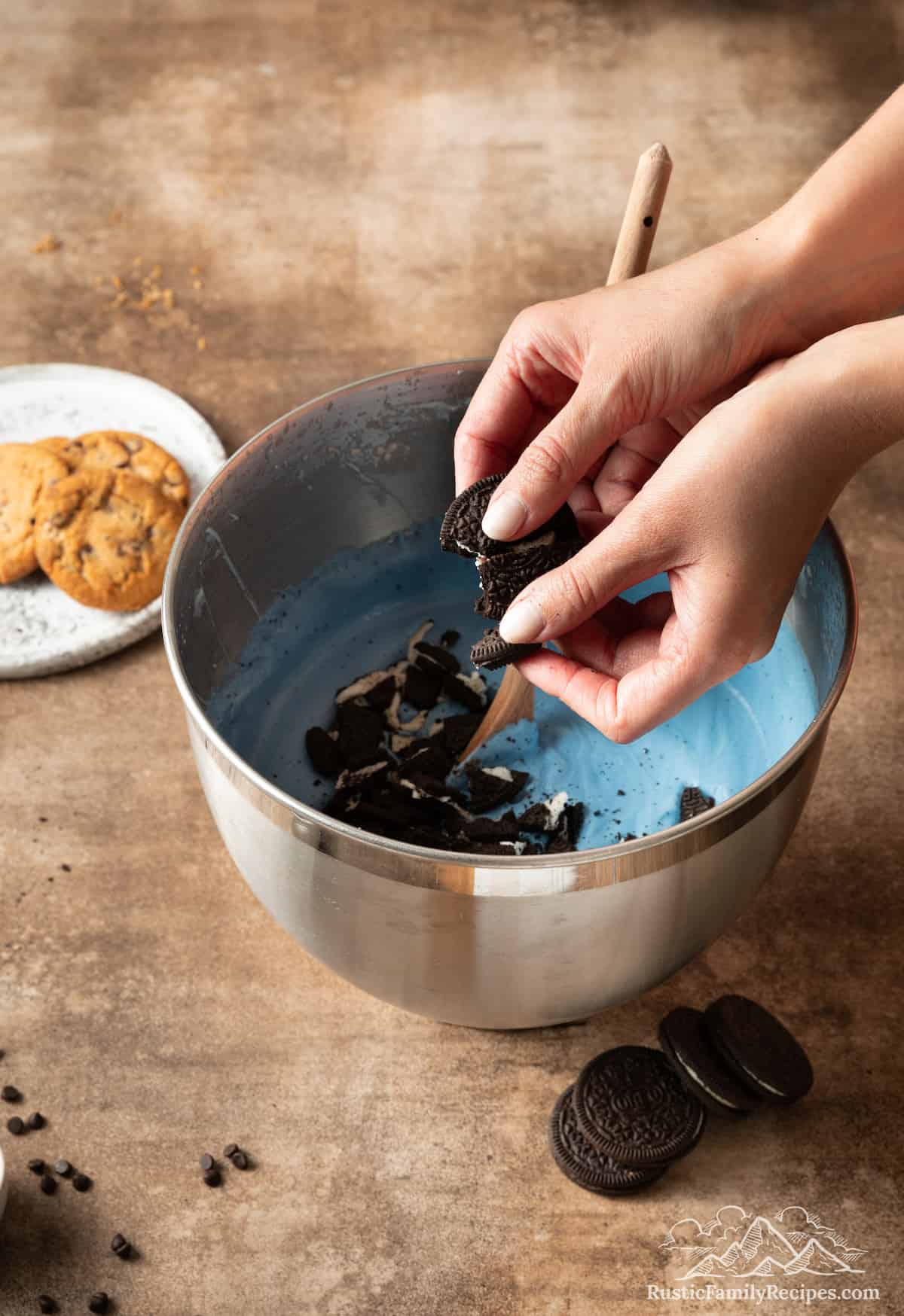 A hand crumbles Oreo cookies into a bowl of blue ice cream.