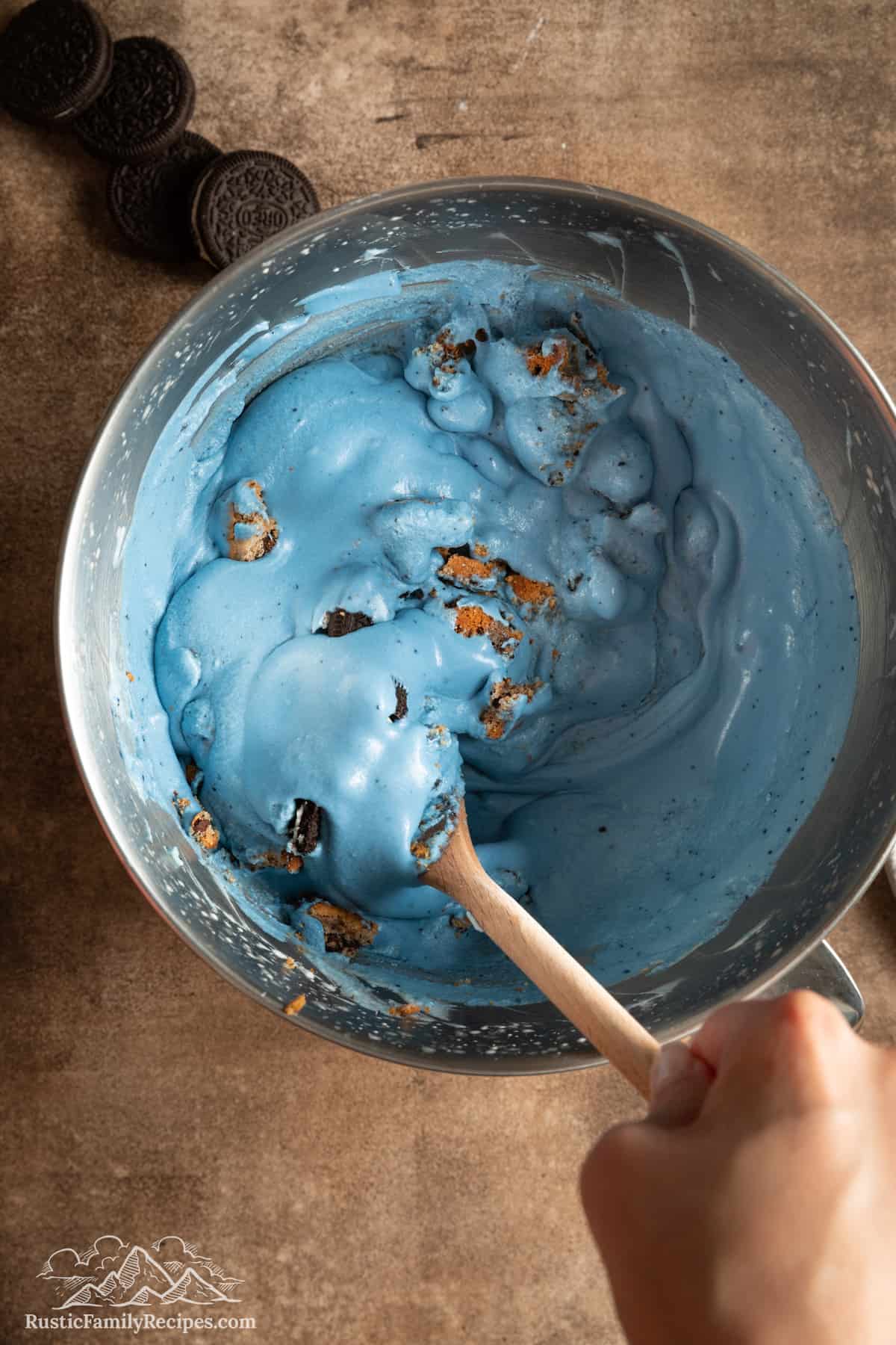 Top view of crumbled chocolate chip cookies and Oreo cookies stirred into blue ice cream with a wooden spoon.