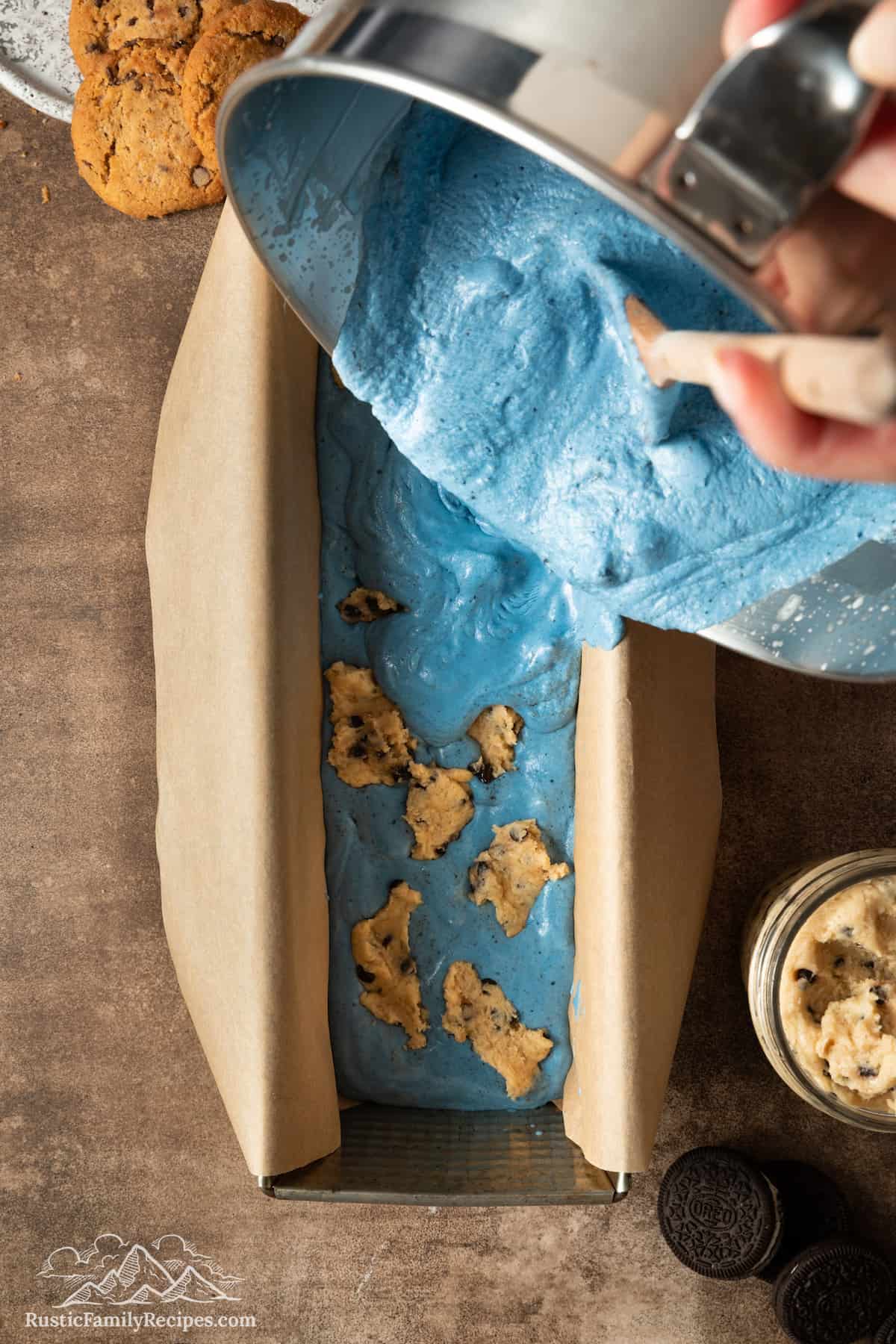 Blue ice cream is spooned over a layer of ice cream and cookie dough in a baking pan.
