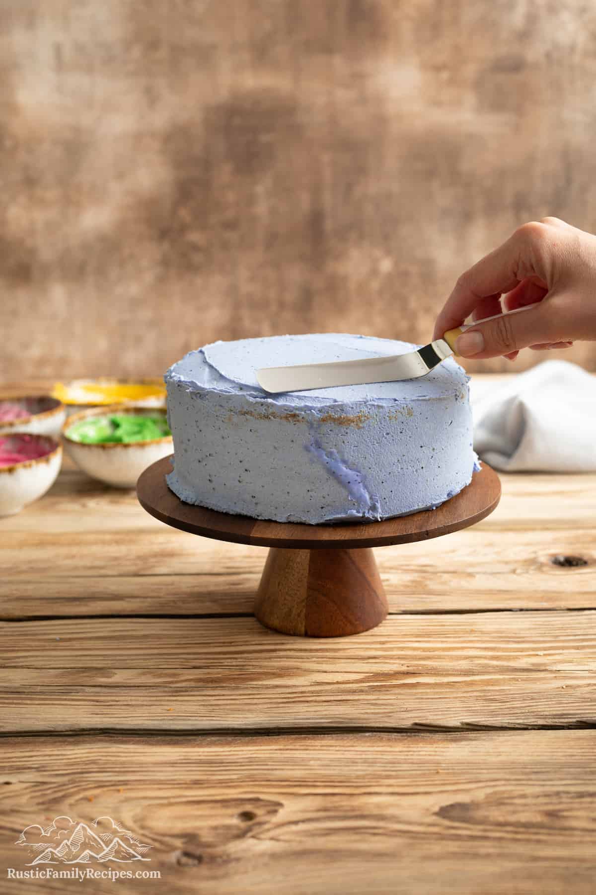 A hand uses an offset spatula to spread blue buttercream frosting over a double-layer cake.