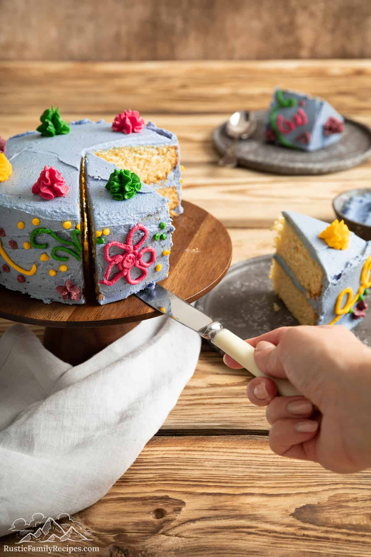 A hand uses a cake knife to lift a slice away from a whole Encanto cake on a cake stand.
