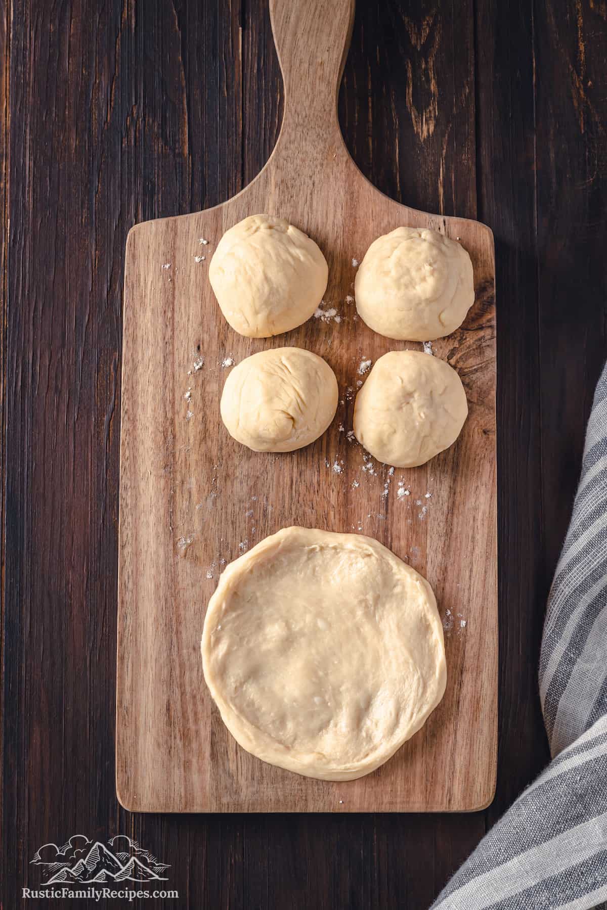 Top view of a flattened round of dough on a wooden cutting board, next to four balls of dough.