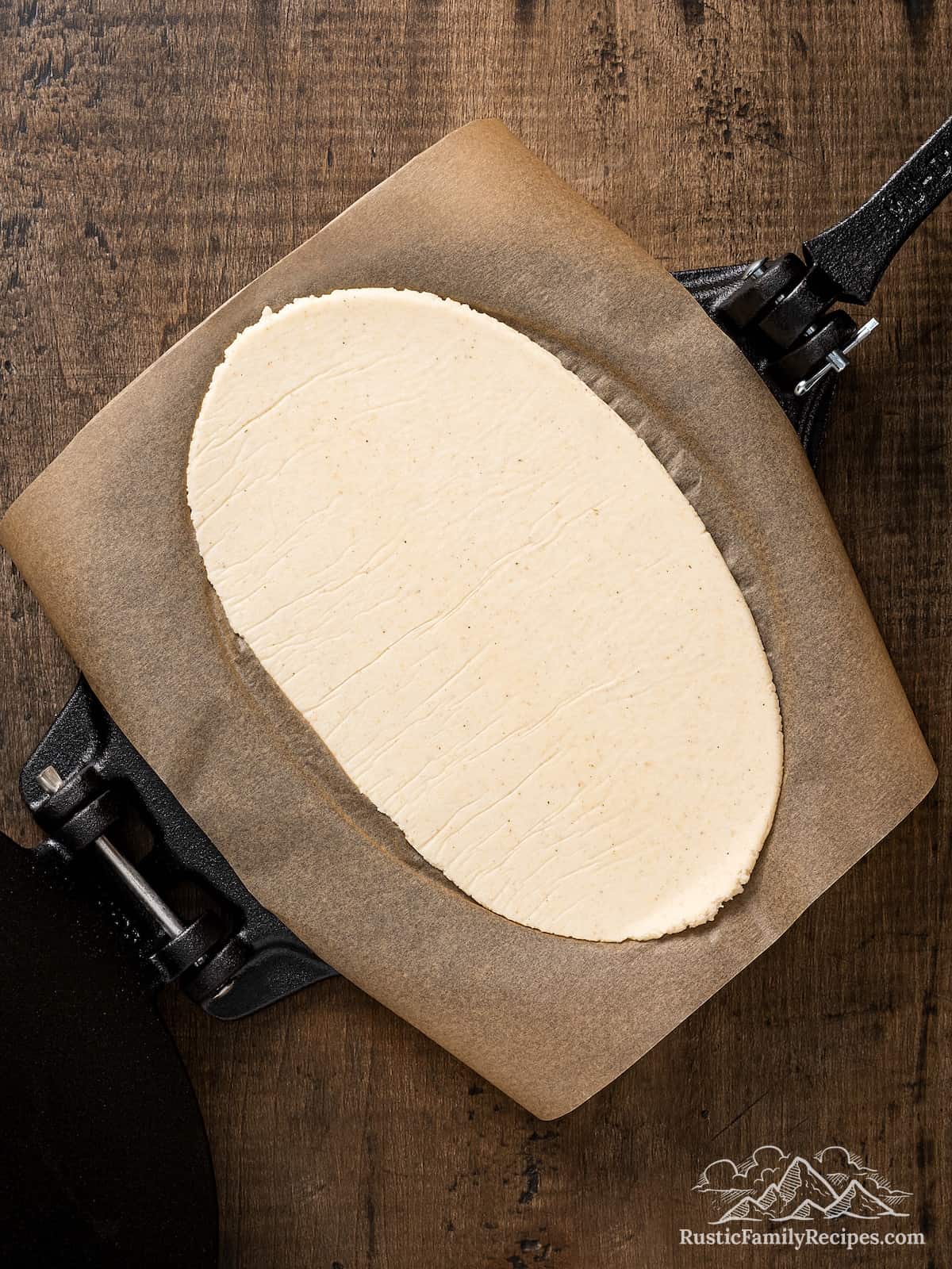 A flattened oval of masa dough on a tortilla press lined with parchment paper.