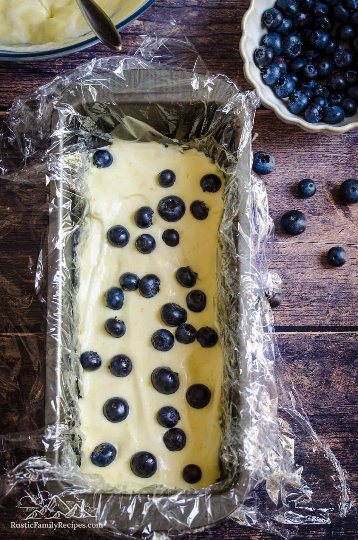 Lemon semifreddo mixture in a rectangular loaf pan lined with plastic wrap, topped with a layer of fresh blueberries.