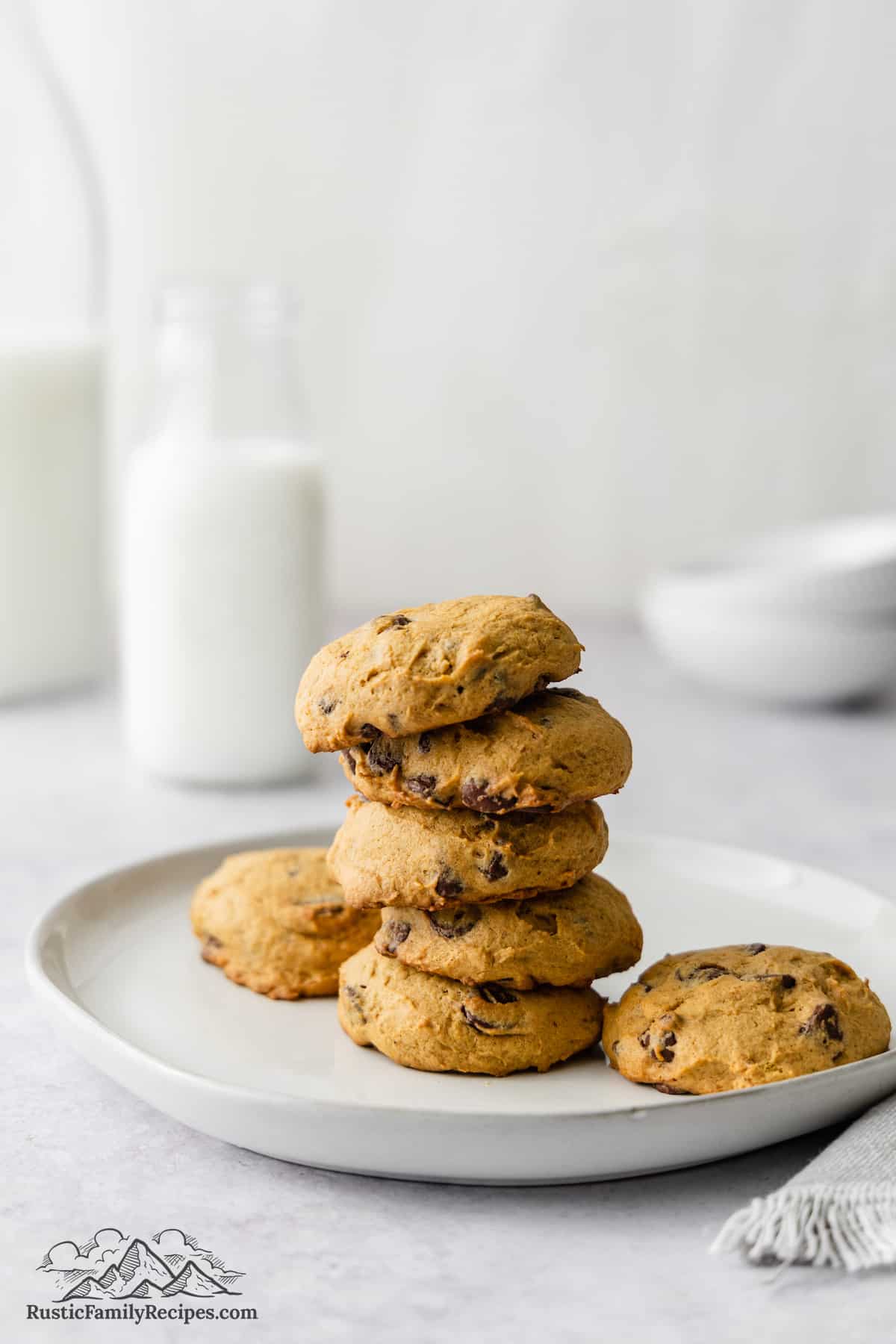 Pumpkin chocolate chip cookies stacked on a plate with a glass of milk in the background.