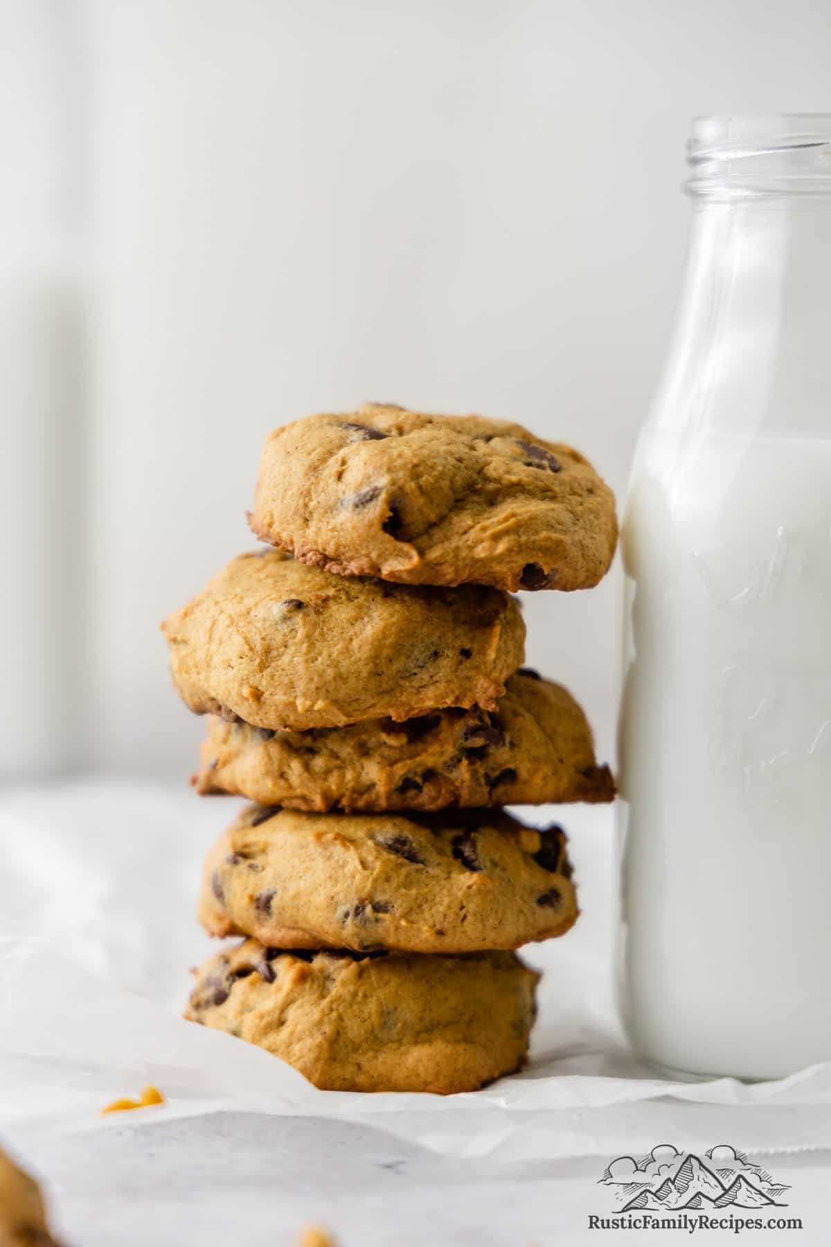 Pumpkin chocolate chip cookies stacked against a glass milk jug filled with milk.