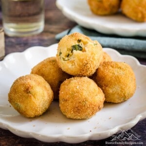 A pile of golden fried arancini with zucchini and goat cheese on a white plate, with a green salad and more arancini in the background.