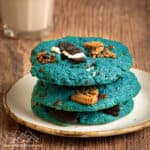 Three Cookie Monster cookies stacked on a white plate, with a glass of milk in the background.