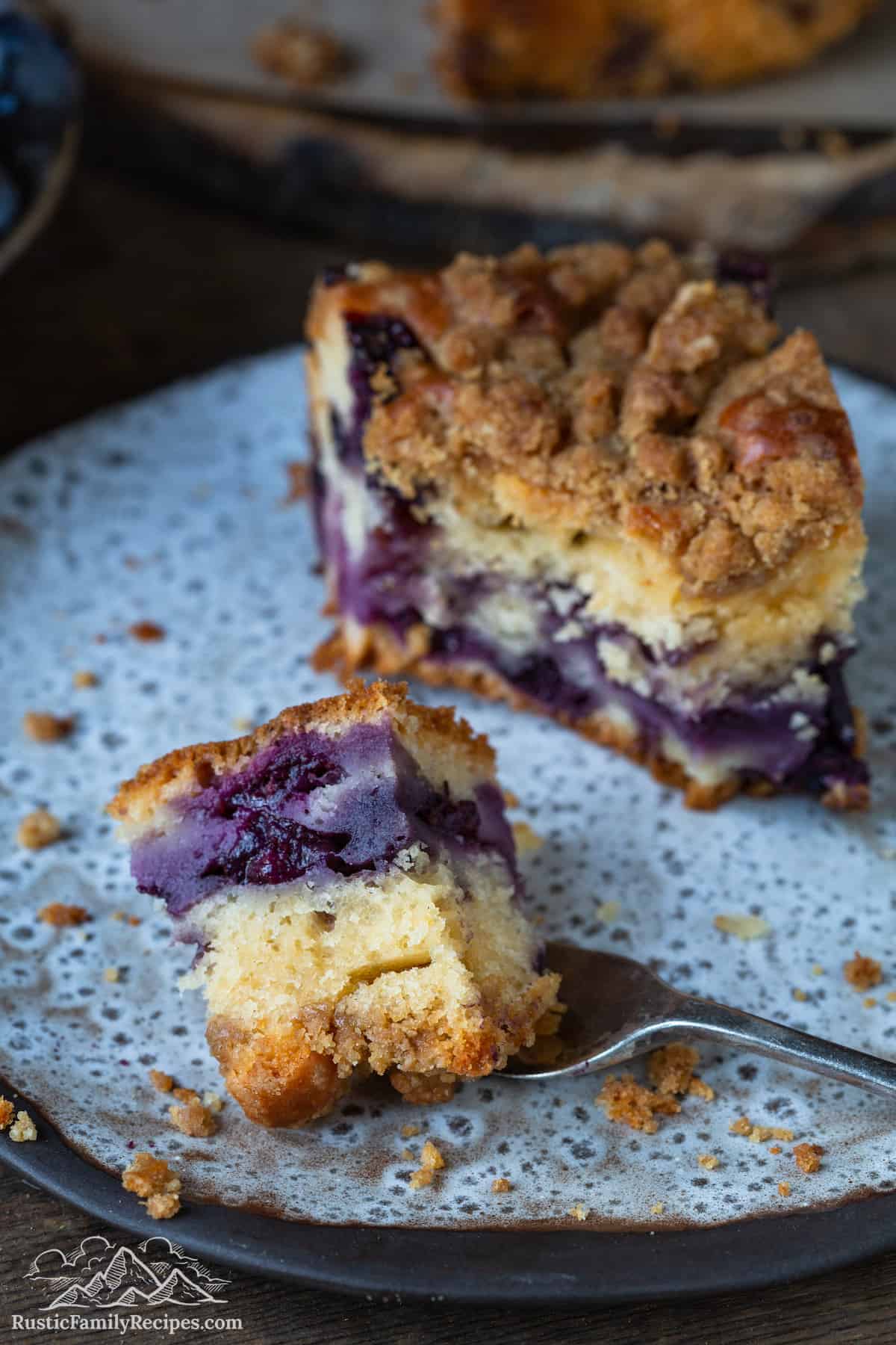 A forkful of blueberry buckle on a plate next to a slice.