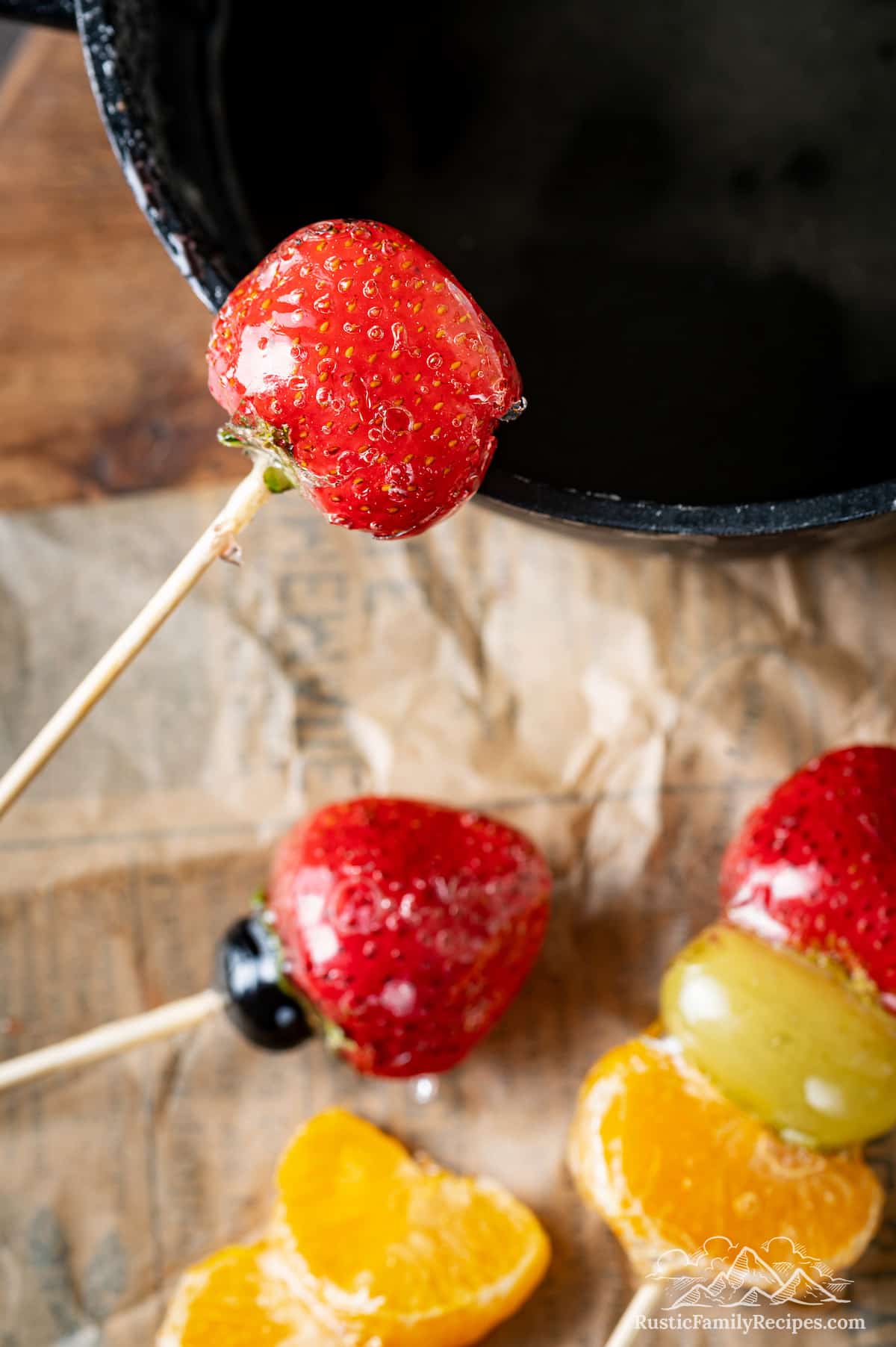 Close up of a candied strawberry on a skewer above a pot, with more tanghulu laying below on parchment paper.