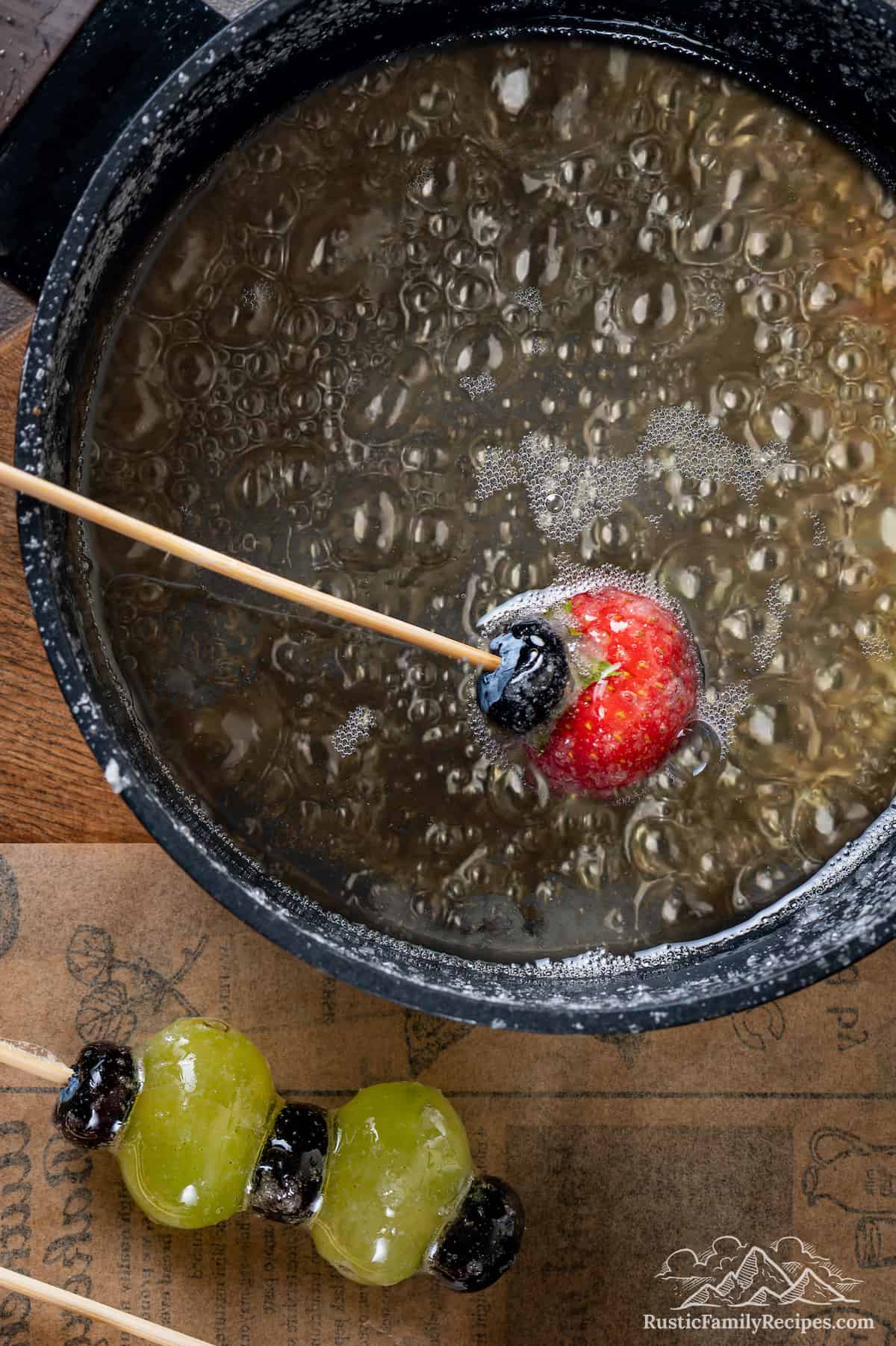 A fruite skewer is dipped into a pot of boiling sugar syrup.