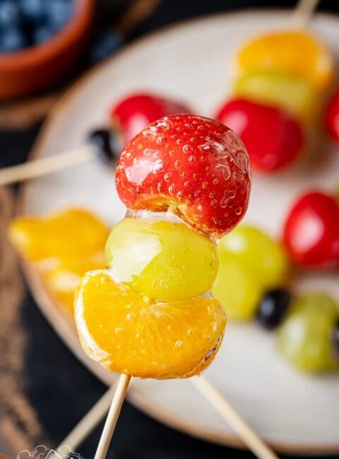 Close up of tanghulu with more fruit skewers on a plate in the background.