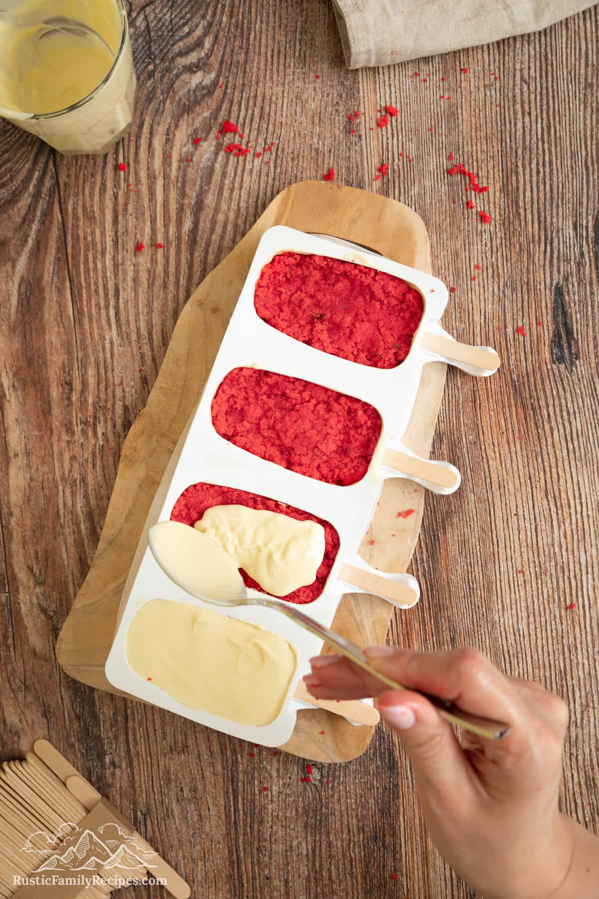 White chocolate is spread over red velvet cakesicles in molds.