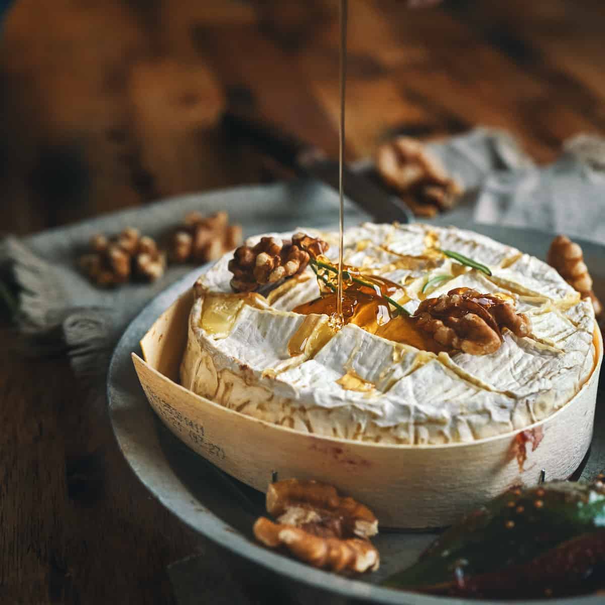 Baked camembert cheese on a plate with walnuts on top, honey being drizzled onto it.