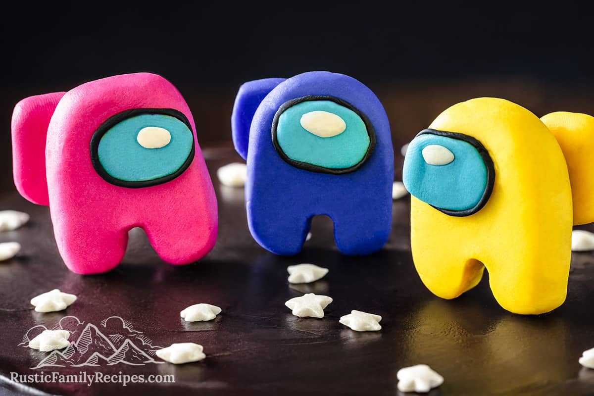 Close up of pink, blue and yellow among us characters made of fondant on a black frosted cake