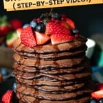 Stack of chocolate chip pancakes with fruit on top, syrup being poured.