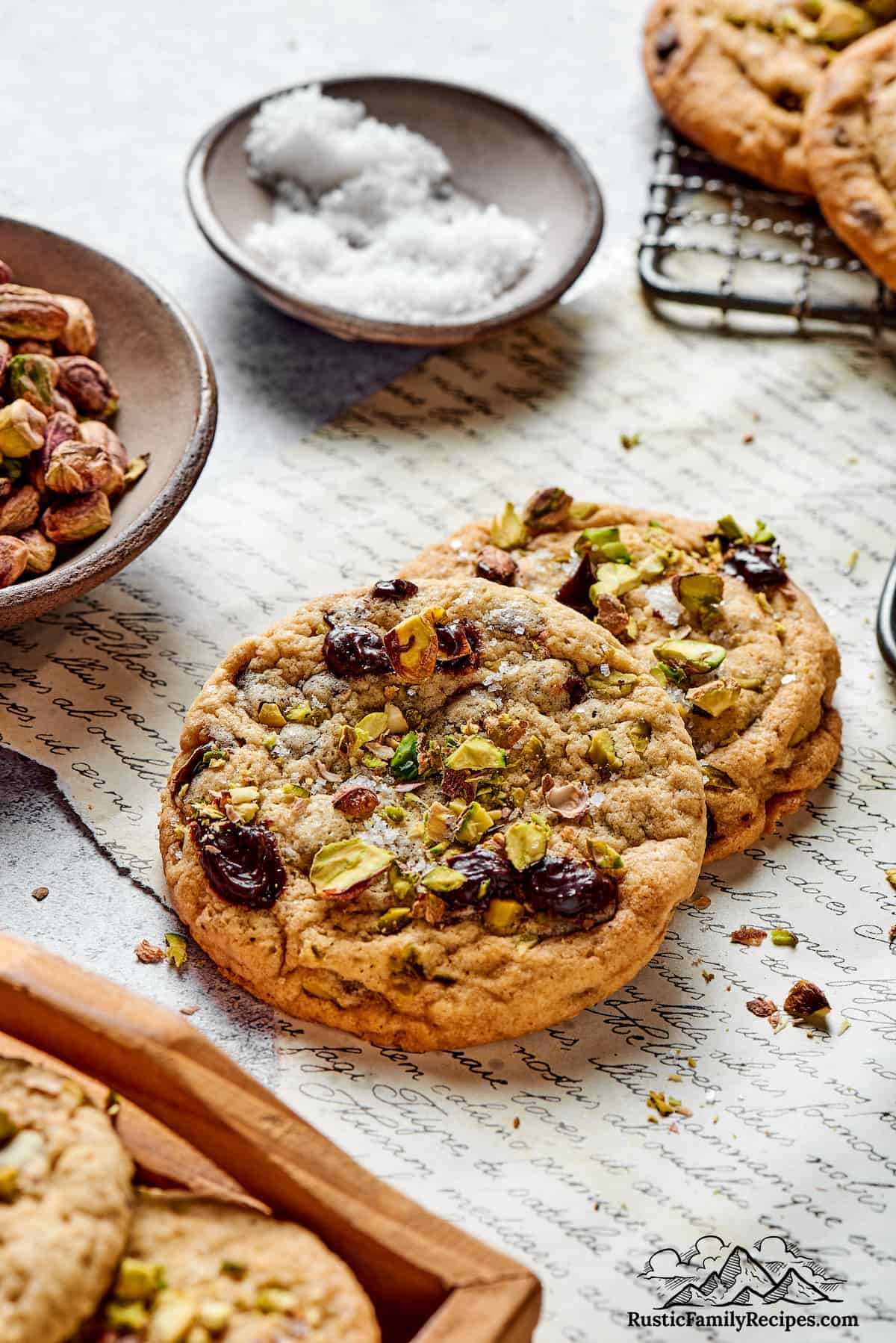 Two cookies on decorative parchment paper with bowls of pistachios and salt
