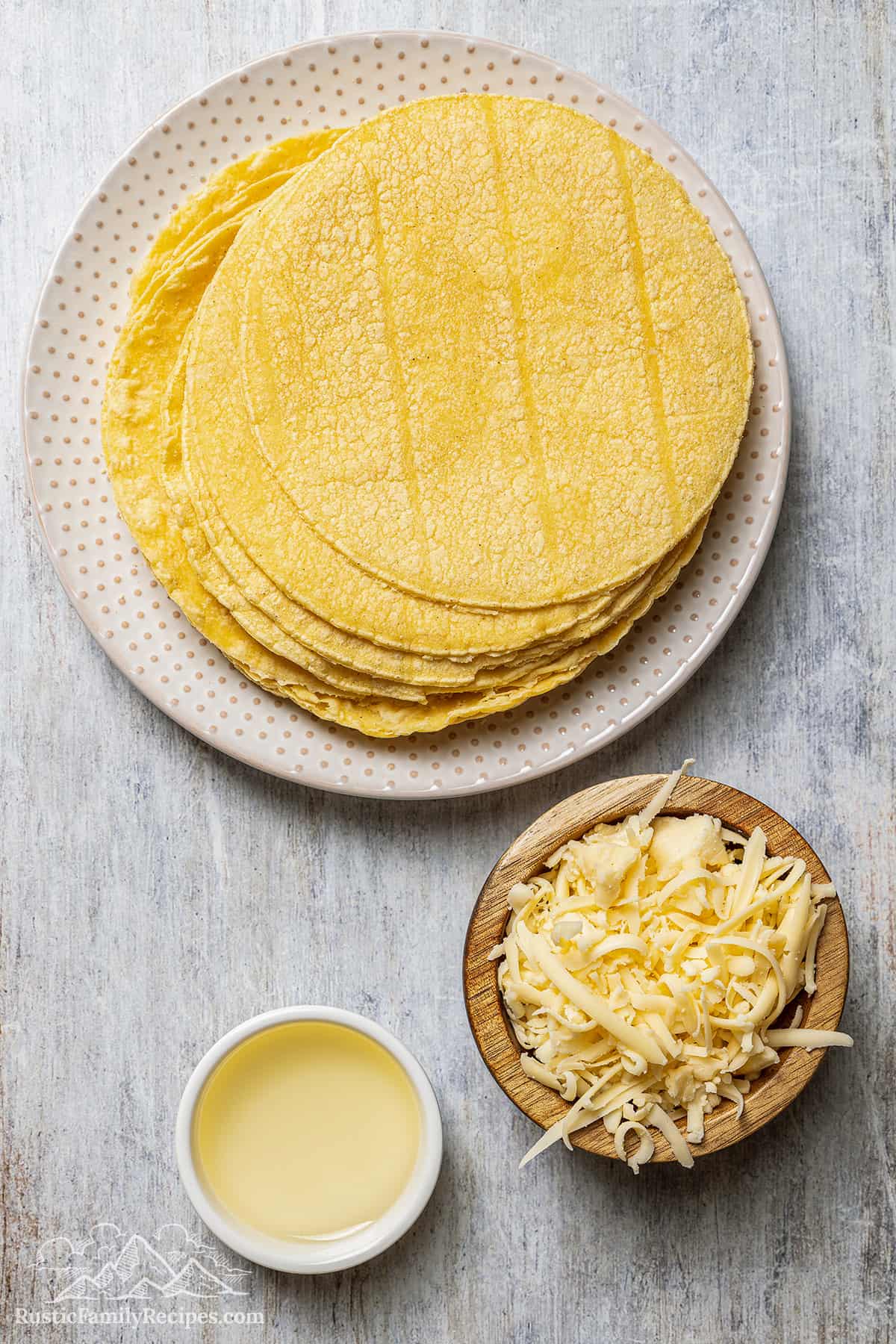 Corn tortillas on a plate next to a small bowl of shredded cheese and oil.