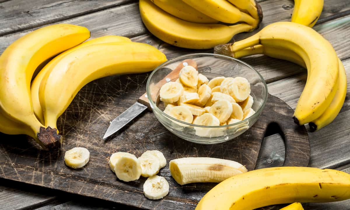 Bananas and banana slices in a plate on a black chopping Board with a knife. On a wooden background.