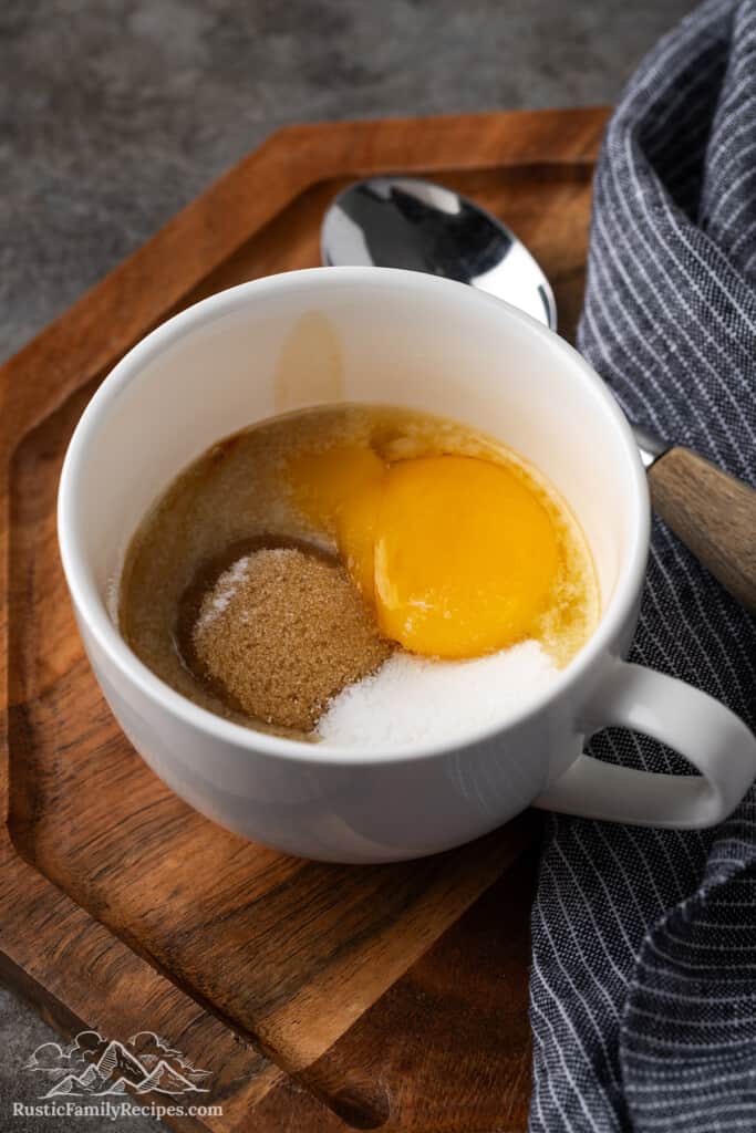 Eggs, sugar, flour and melted butter in a white mug.