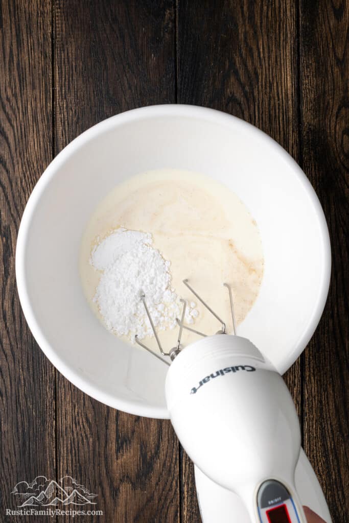 A hand mixer is used to combine the ingredients for whipped cream in a white mixing bowl.