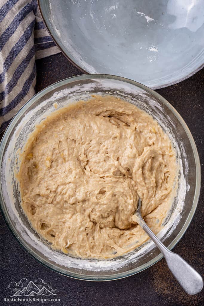 Banana bread muffin batter in a large glass bowl with a stirring spoon.