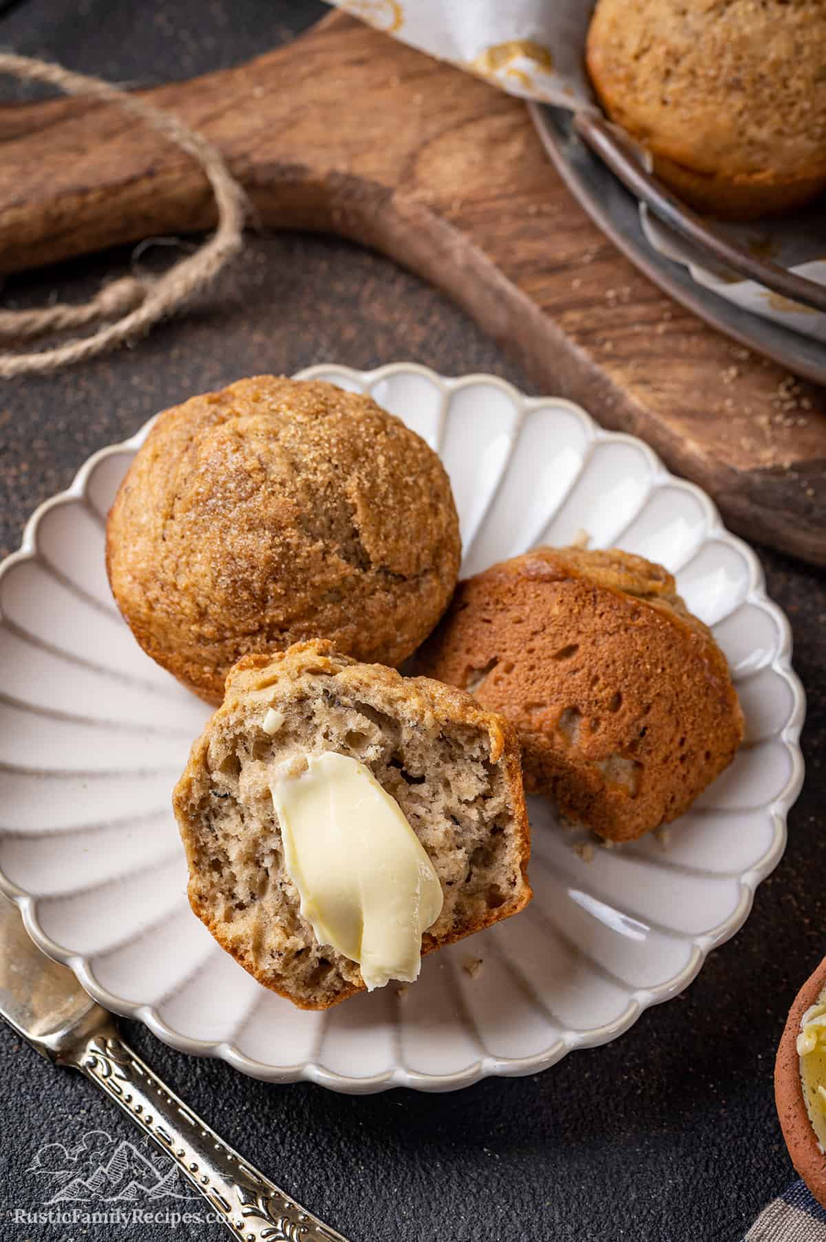 One half of a banana bread muffin spread with butter on a plate next to its other half, and a whole banana muffin.