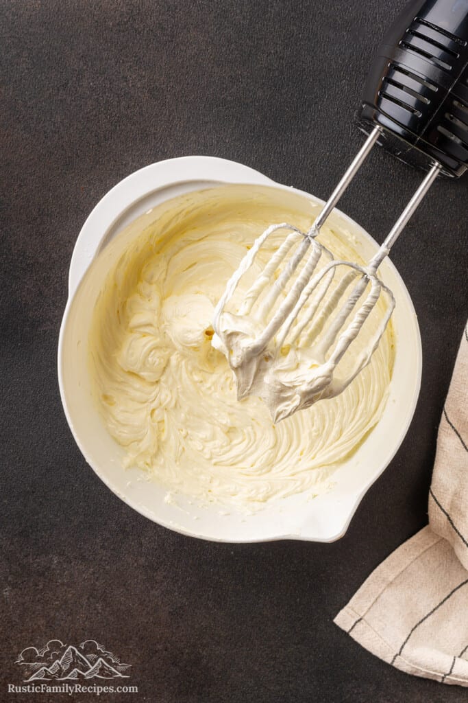 The whisk attachment of a hand mixer resting on the edge of a bowl of buttercream frosting.