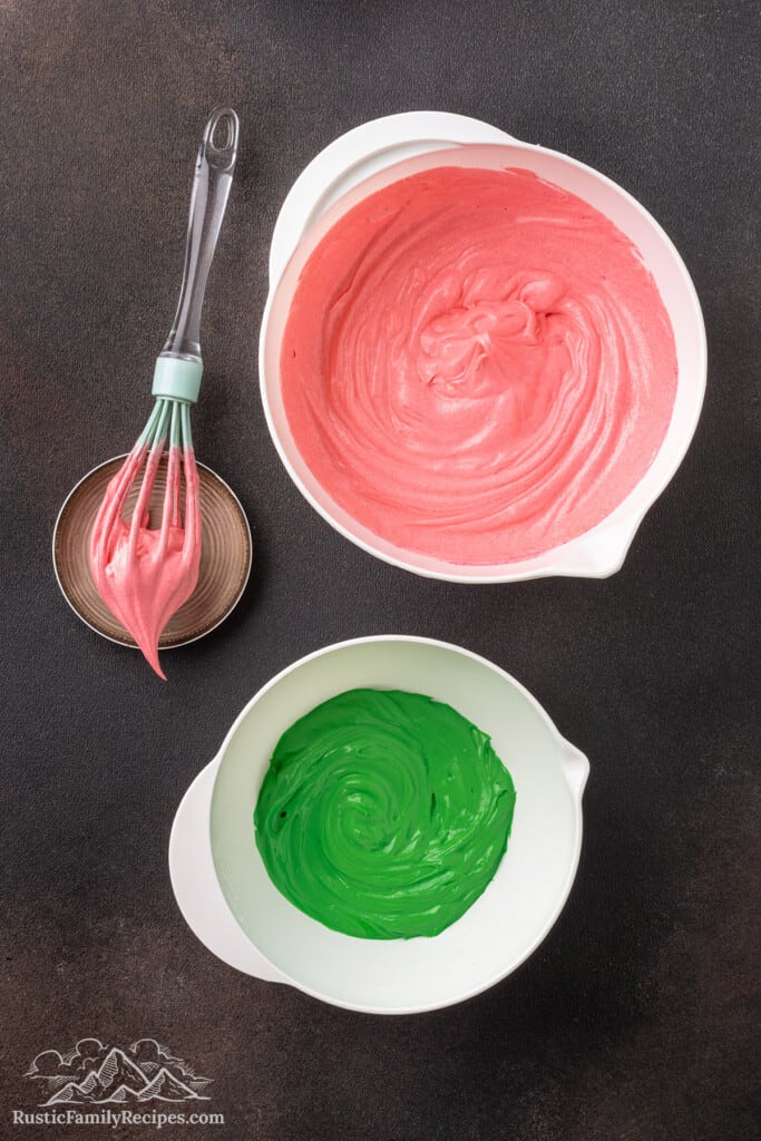 A bowl of pink frosting next to a second bowl of green frosting, next to a whisk.