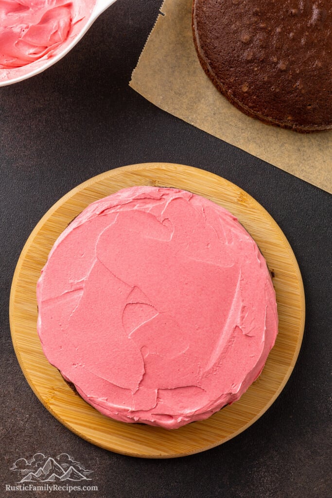 Overhead view of pink frosting spread over the first chocolate cake layer on a wooden cake stand.