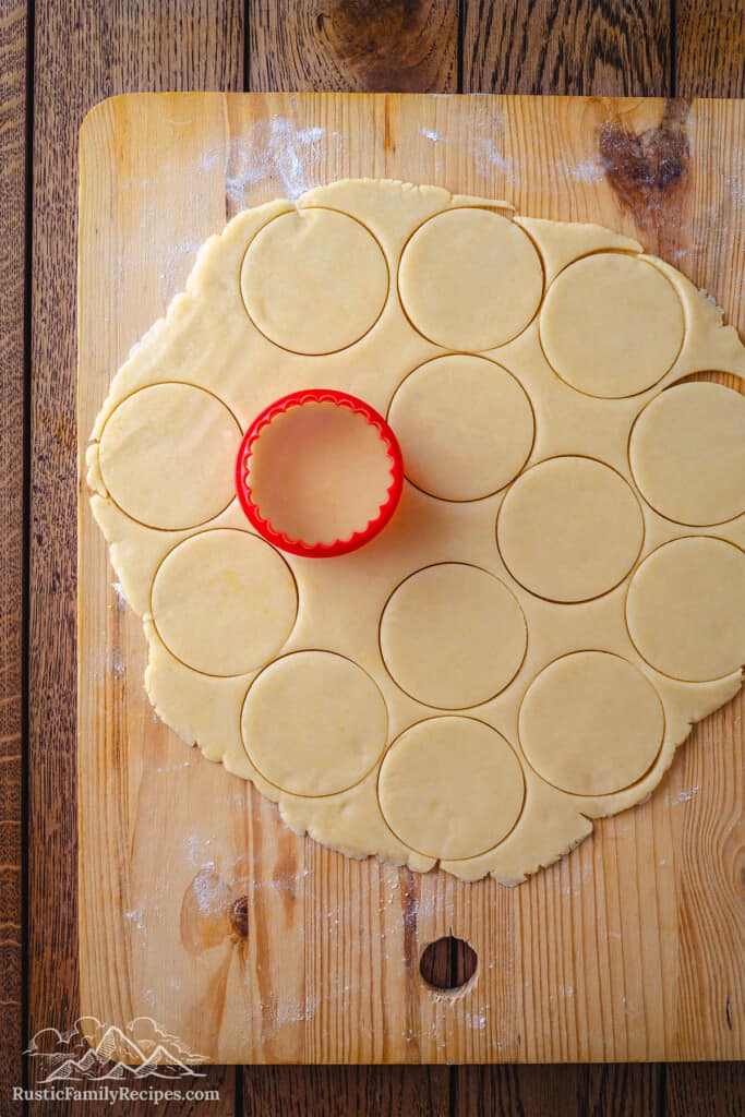 A round cookie cutter is used to cut out rounds of pizzicati cookie dough on a large round cutting board.