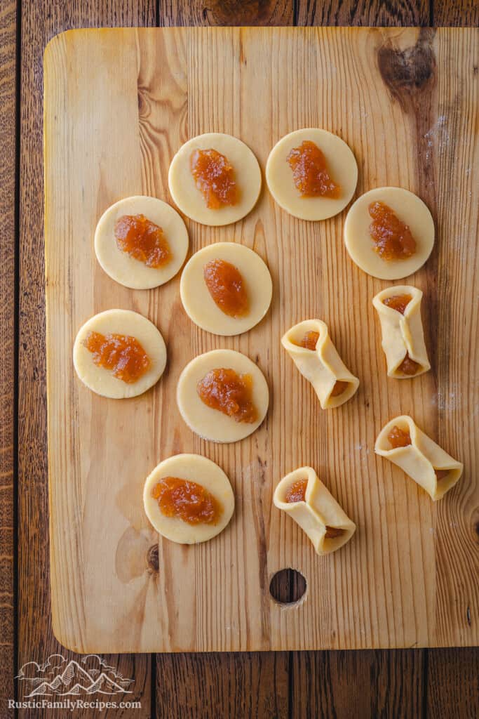 Overhead view of small dough rounds topped with jam being folded into pizzicati cookies on a large wooden cutting board.
