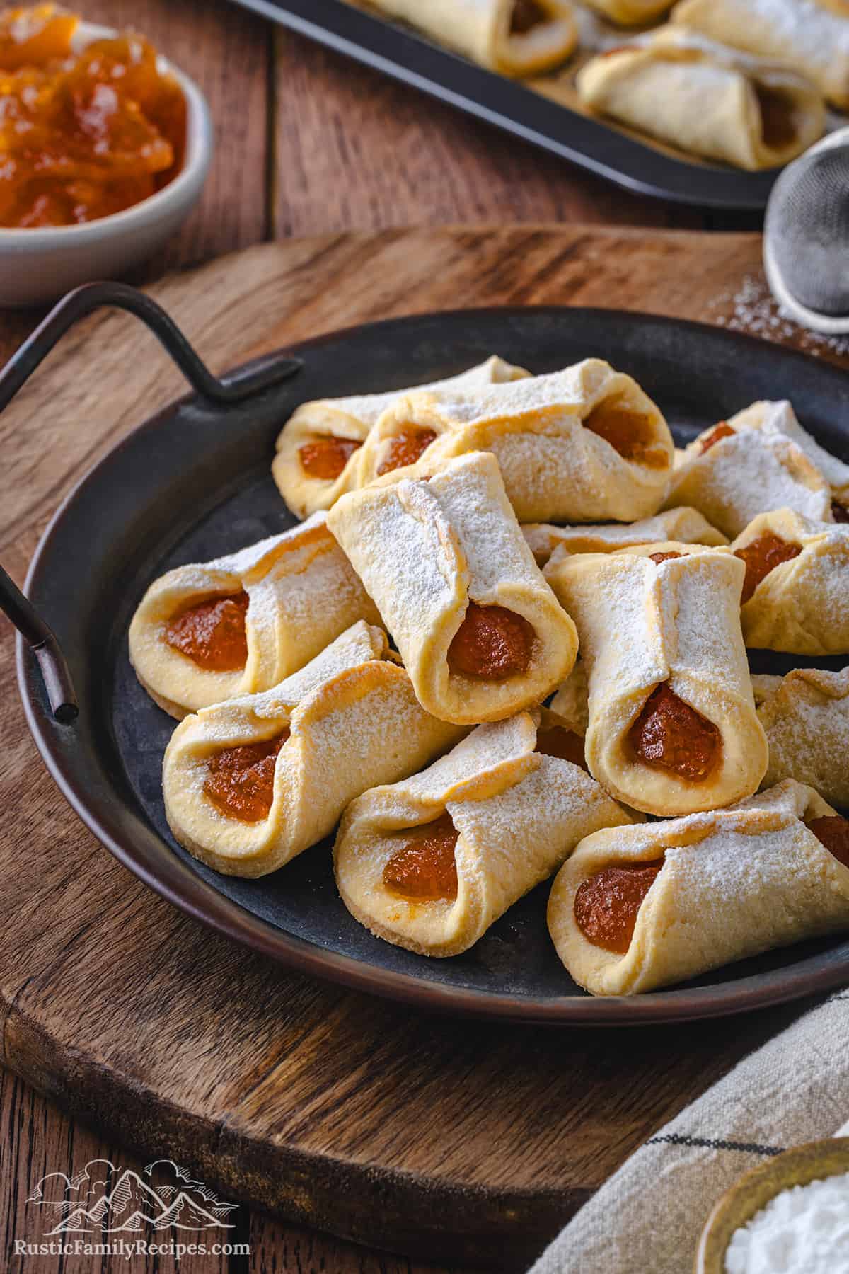 Close up of assorted pizzicati cookies filled with jam and dusted with powdered sugar on a dark plate, with a bowl of apricot jam and more cookies in the background.